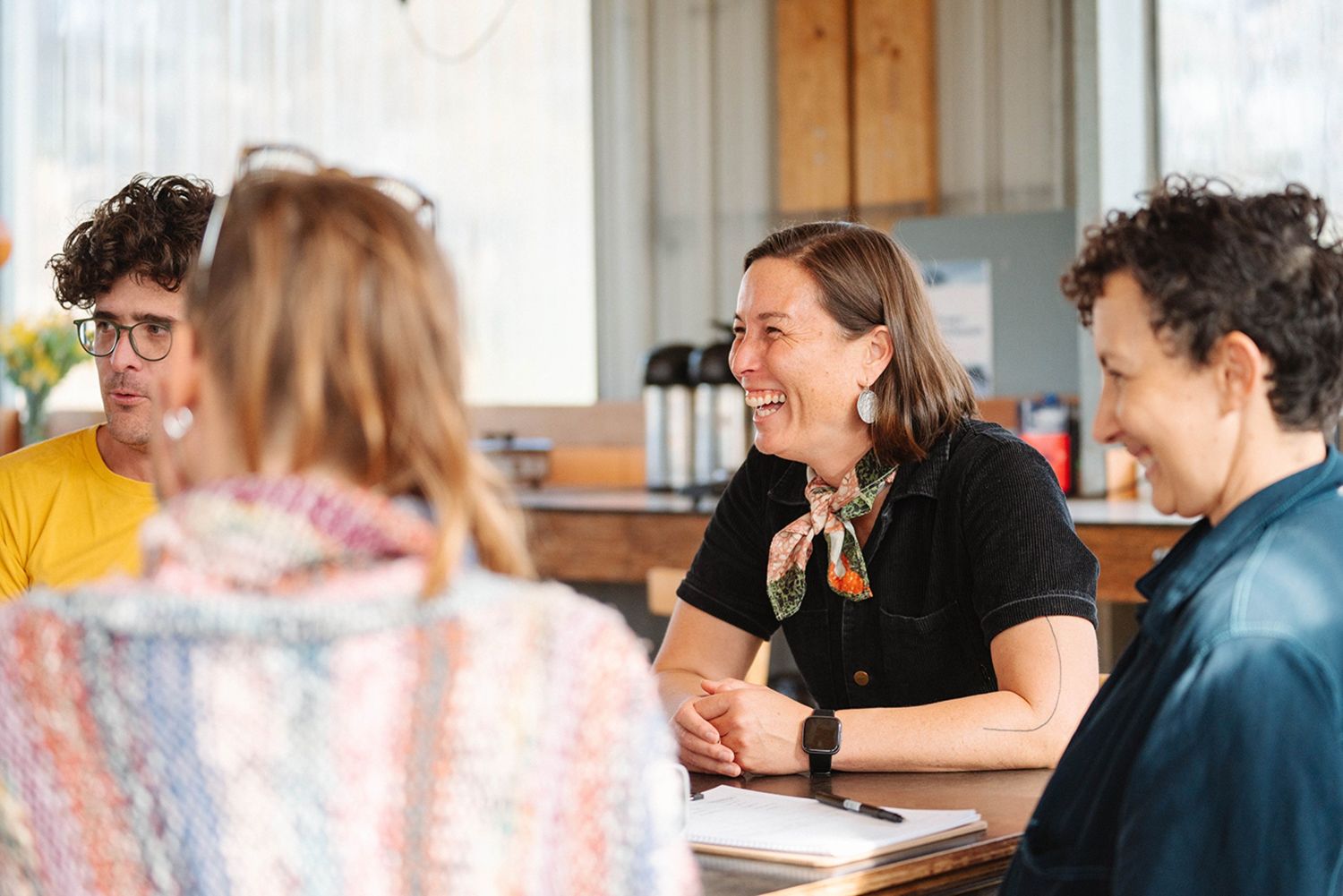 A group of young people sit around a table, with the attention focused on someone off camera, all smiling happily
